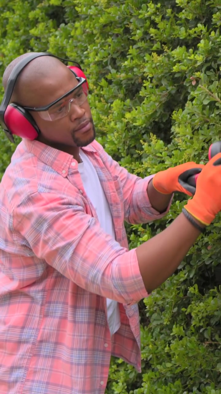 Vertical video: Starting man in plaid and orange gloves raising red trimmer in yard, shaping hedge