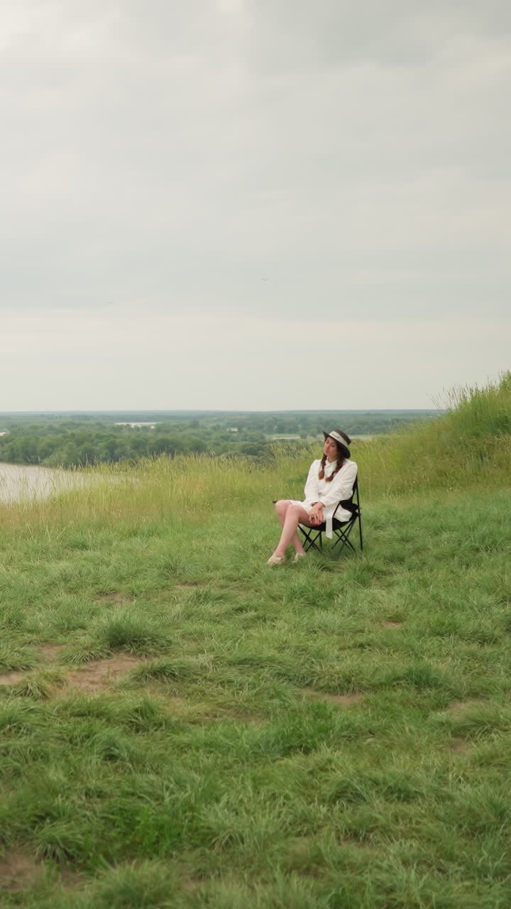 한 화가, 모자와 체크 셔츠를 입고, 구름이 은 하늘 아래 울창한 잔디 한가운데의 보드에 그림을 그리는 데 집중하고 있습니다. 모자와  드레스를 입은 여자가 의자에 편안하게 앉아 있습니다.