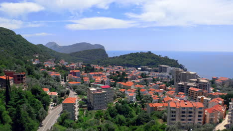Die-Küstenstadt-Petrovac,-Montenegro,-An-Der-Adriaküste,-Mit-Häusern-Mit-Roten-Ziegeldächern,-Wolkenkratzern,-Umgeben-Von-Küstenklippen-Und-Wäldern,-Sonniger-Tag,-Blauer-Himmel-Mit-Wolken-Darüber