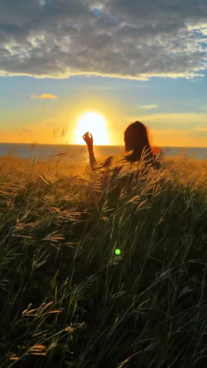 Silhueta de Mulher na Grama ao Pôr do Sol. Silhueta de mulher sentada em grama costeira alta de frente para um pôr do sol brilhante sobre o oceano, criando uma atmosfera emocional calma