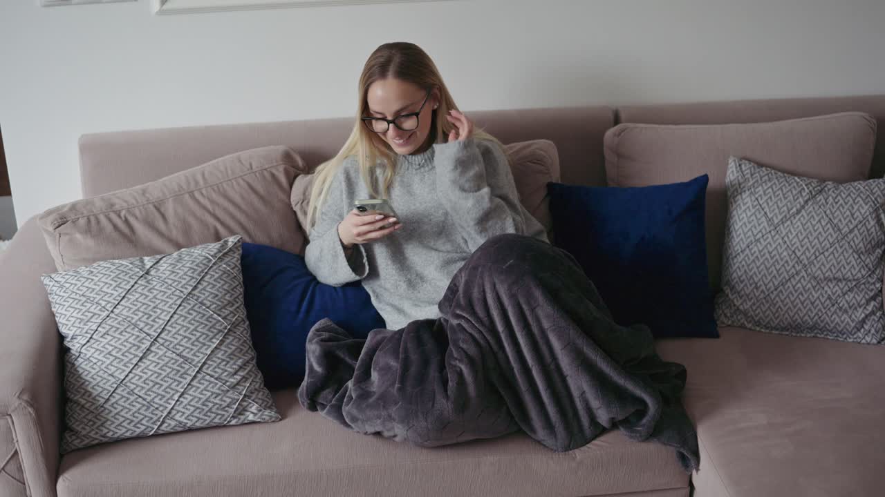 Una giovane donna con gli occhiali è seduta comodamente su un divano a casa, sorridente mentre usa il suo smartphone. Avvolta in una morbida coperta e circondata da cuscini decorativi.