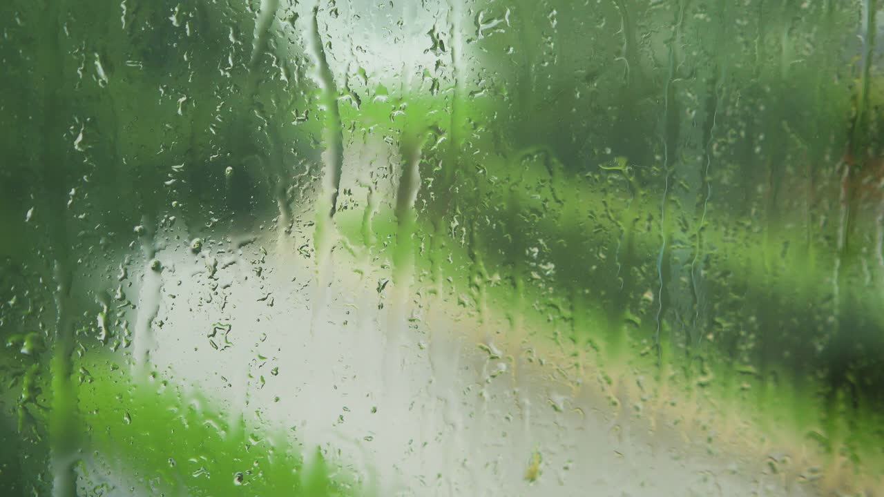 Rain drops sliding slow on window glass in rainy day, de-focused street with moving car in background, medium closeup shot