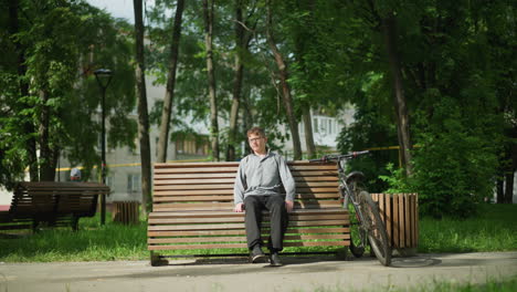 boy wearing glasses, gray top sits on wooden bench in park, stretches forward while adjusting himself, places right foot over left leg, bicycle parked nearby, greenery and trees around