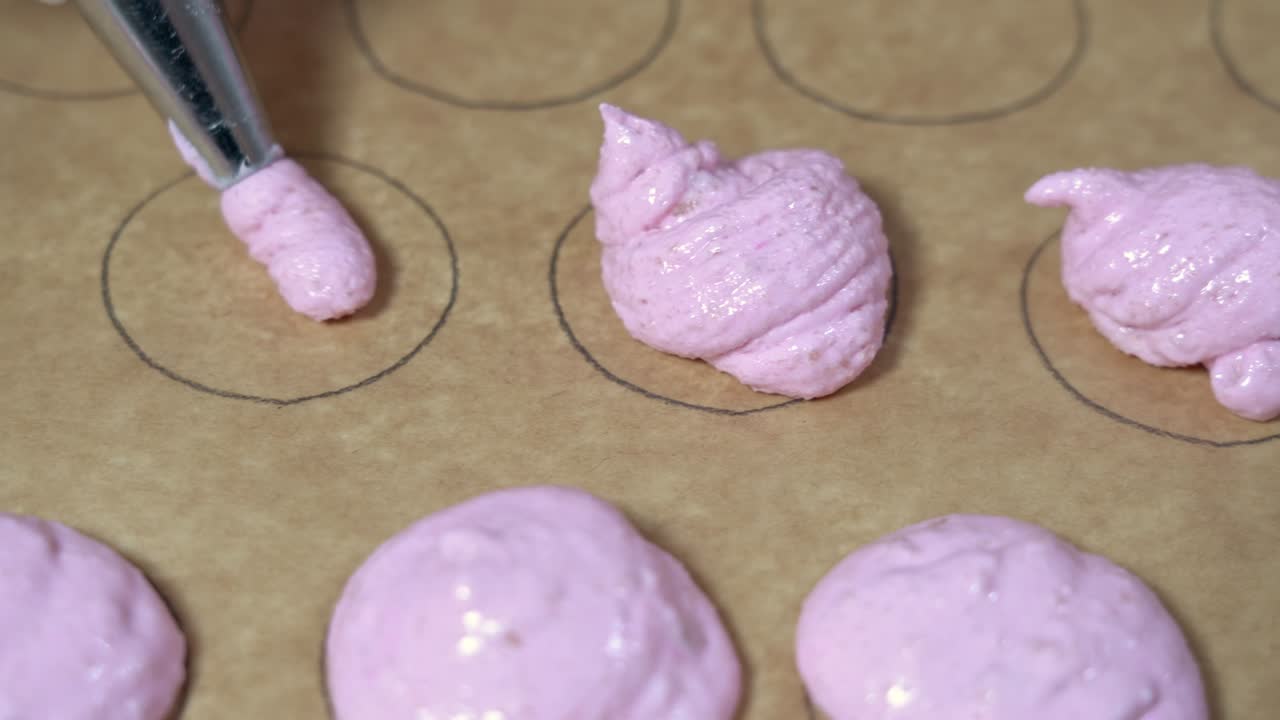 una toma de cerca de un panadero usando una bolsa de pastelería con masa rosa de macarrón para llenar formas circulares en papel de pergamino.