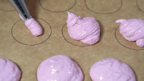 close up shot of baker using a pastry bag with macaron pink batter to fill up circular shapes on parchment paper.