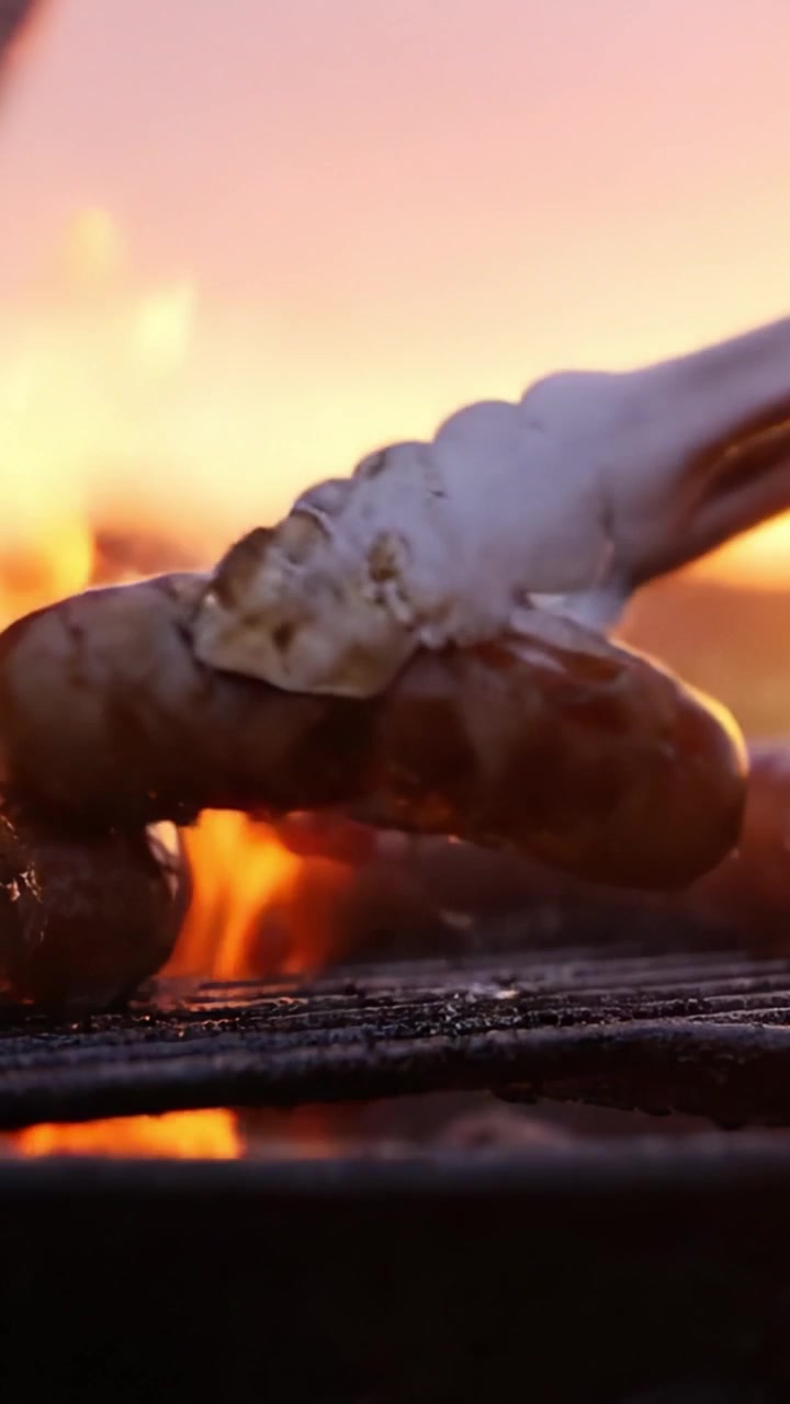 Un primo piano di salsicce sfrigolanti che grigliano su una fiamma viva al tramonto, mettendo in mostra la deliziosa doratura e la consistenza succosa della carne alla brace.