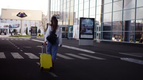Stylish-woman-walk-with-trolley-yellow-case-by-empty-airport-terminal-outside-road,-slender-female-wearing-jeans-and-brown-boots
