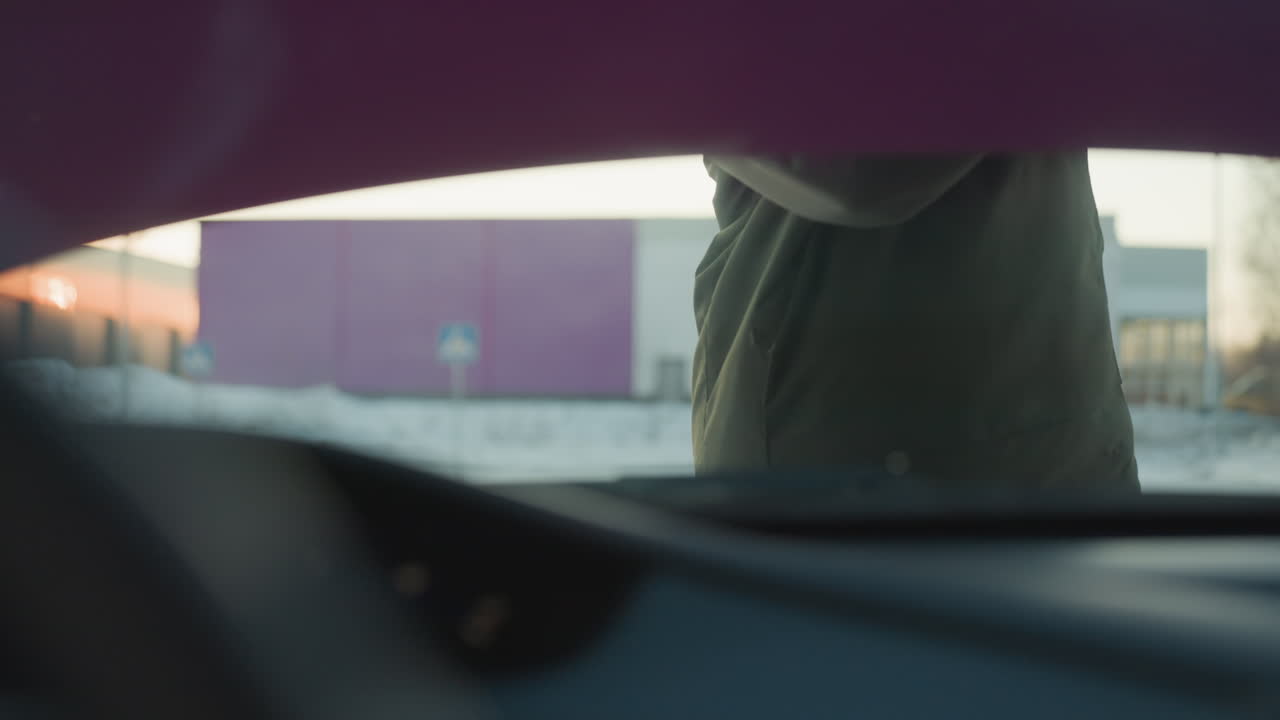 Close up from inside car shows person opening bonnet and placing rod support to hold it up for inspection, with winter background and distant buildings visible in cold outdoor maintenance environment