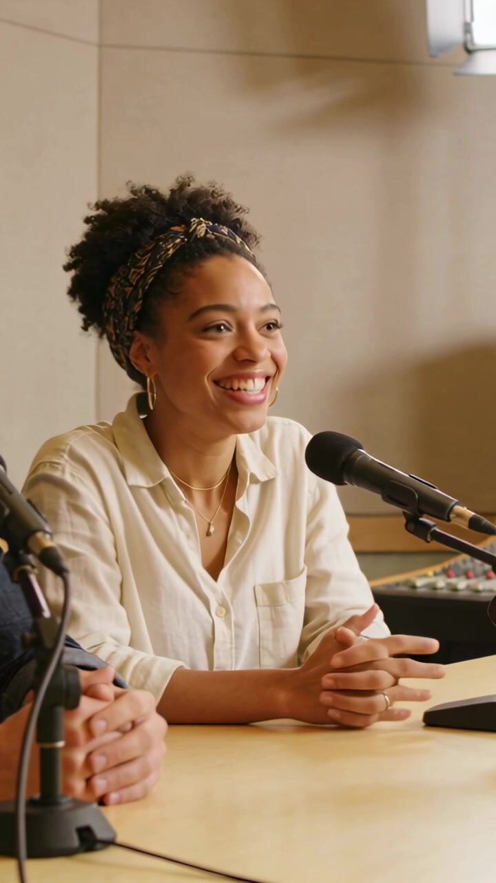 Vertical video: Smiling host replying to cohost in studio, wearing patterned headband, with mics
