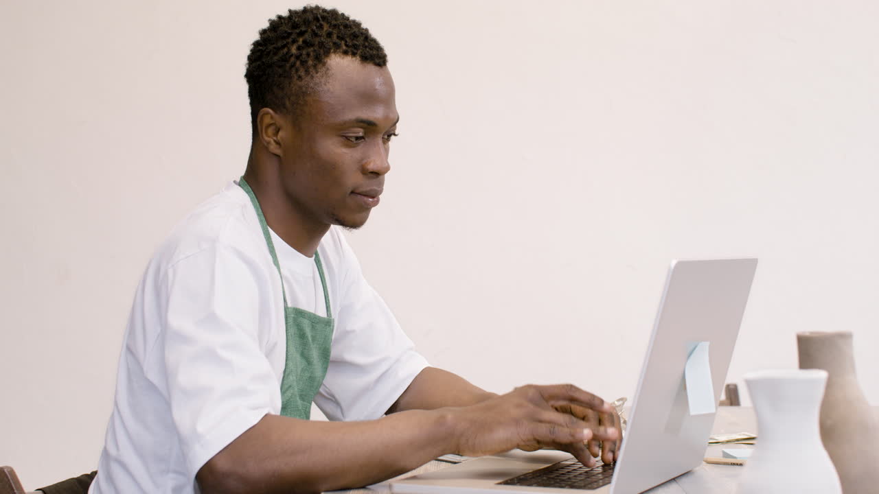 American Clerk In Apron Sitting In Front Of Laptop And Typing On Keyboard