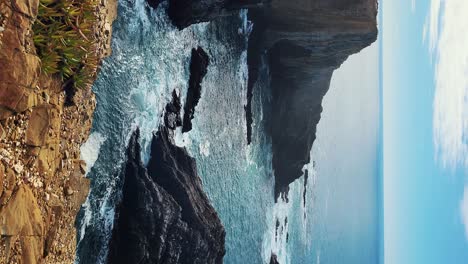 Zambujeira-do-Mar-over-the-sea-shore-with-ocean-waves,-cliffs-and-sand-dunes-covered-by-green-vegetation-red-leaves-of-sour-fig,-sunny-day,-clear-blue-sky