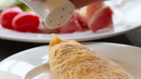 a close-up of seasoning being added to an omelette, accompanied by tomatoes and bacon on a plate.