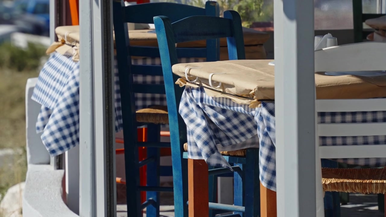 Colorful wooden chairs and blue checkered tablecloths line a sunny outdoor cafe terrace in Santorini with no people present.