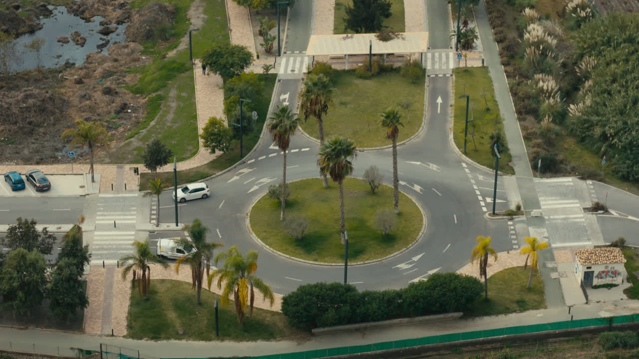 Top-down view of landscaped roundabout with palm trees and cars in motion, surrounded by walkways, roads and mixed vegetation