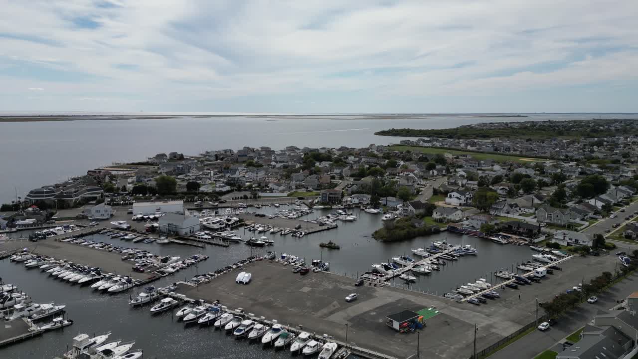 Højt luftfoto, der flyver sidelæns over en naturskøn marina fyldt med fortøjede både og yachter, afslører den tilstødende kystby og rolige bugtvande under en delvist overskyet himmel