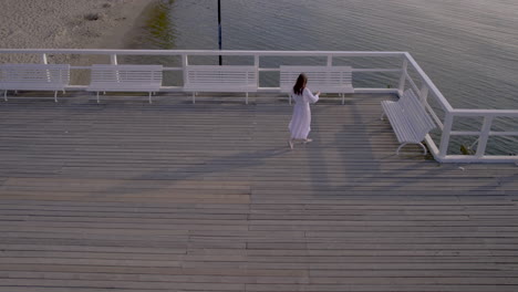 Woman-walks-alone-towards-benches-on-wooden-pier,-long-shadow-cast-on-wood-path