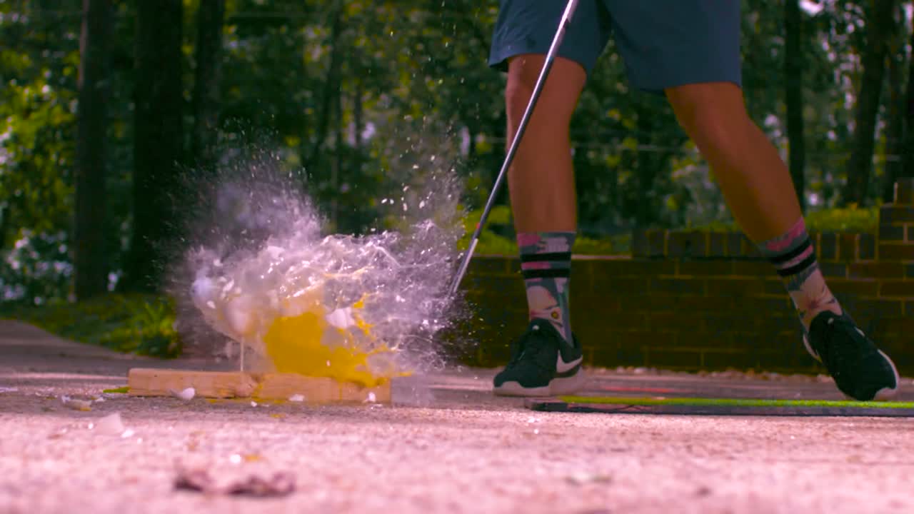 A golf ball shattering and destroying three eggs in slow motion by the golf ball being hit with a golf stick or bat during summer time with sun. The eggs explode their shells revealing insides, yolk.