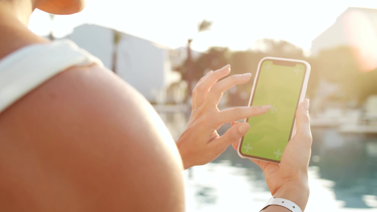 vista por encima del hombro de la joven mujer caucásica que está tomando un bronceado en la piscina y usando un teléfono inteligente con tecla de croma y seguimiento de movimiento. pantalla verde. cerca.