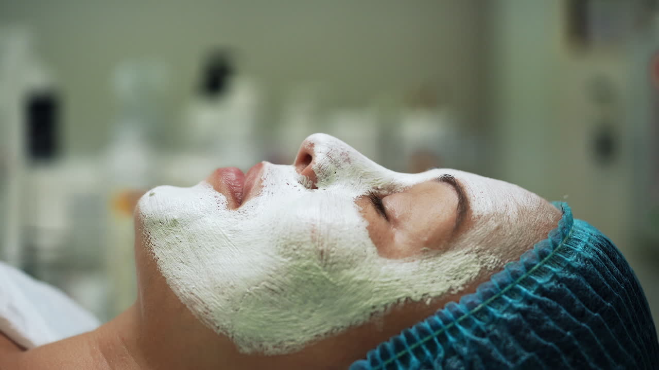 Woman in blue protective cap lies having a white creamy face mask. Beauty procedure in modern salon. Side view close up.