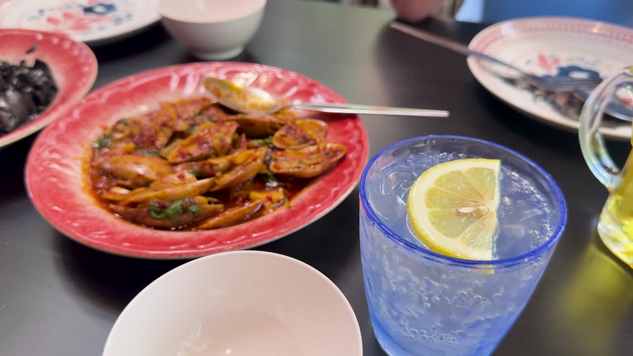 A close-up view of a vibrant Thai seafood dish and a glass of fizzy water with lemon, shot in bright indoor lighting with minimal camera movement