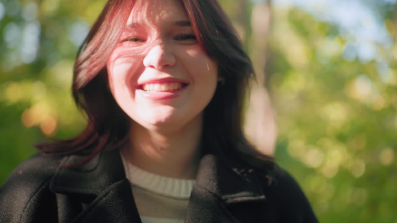 Cheerful woman smiles warmly under sun beam, head rolling in relief, eyes bright, forest bokeh behind, warm autumn light soft on face, cozy coat, joyful mood captured during quiet walk in park