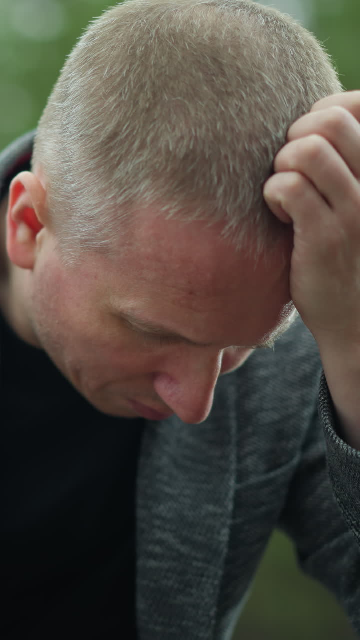 una vista cercana de un hombre con una chaqueta gris y pantalones vaqueros azules sentado en una vía ferroviaria, sosteniendo una pistola con la cabeza inclinada y la mano descansando en su cabeza, el fondo muestra un bosque verde borroso