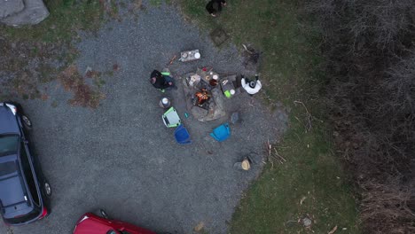 aerial top-down view of a group of people sits around a fire, have a rest in front of country house beside a red car, shot by drone flying up on an autumn day in rural rome, pennsylvania