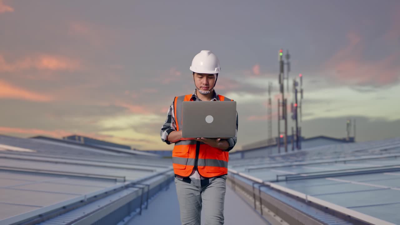 Front View of Asian Male Engineer Use Computer While Walking at Industrial Rooftop Solar Panels