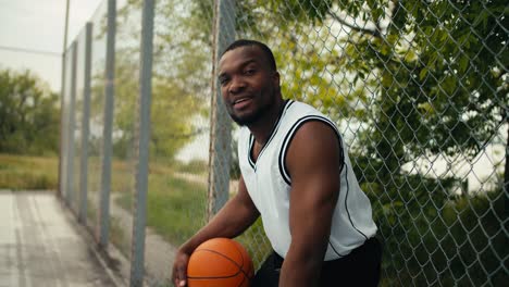 tired black man in a white t-shirt holding a basketball in hands and posing on the sports ground outdoor