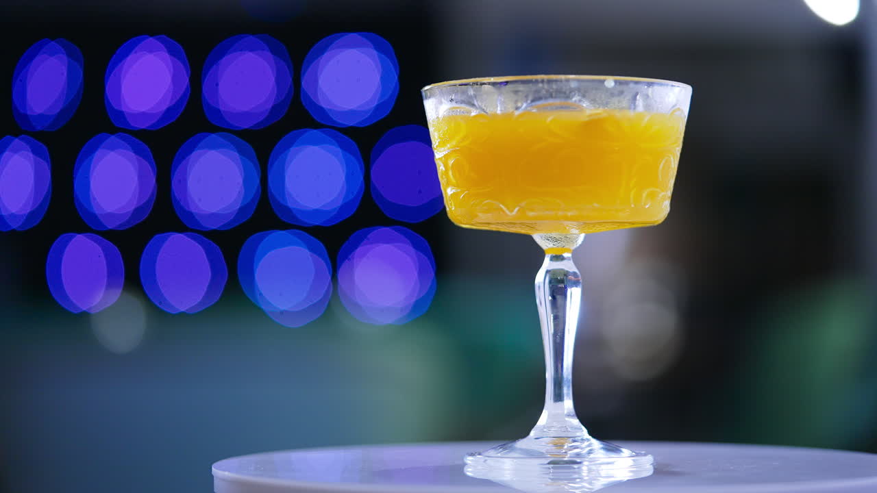 White rotating table with a glass of cocktail. Wide wineglass filled with orange beverage. Close up. Blue lights at backdrop in blur.