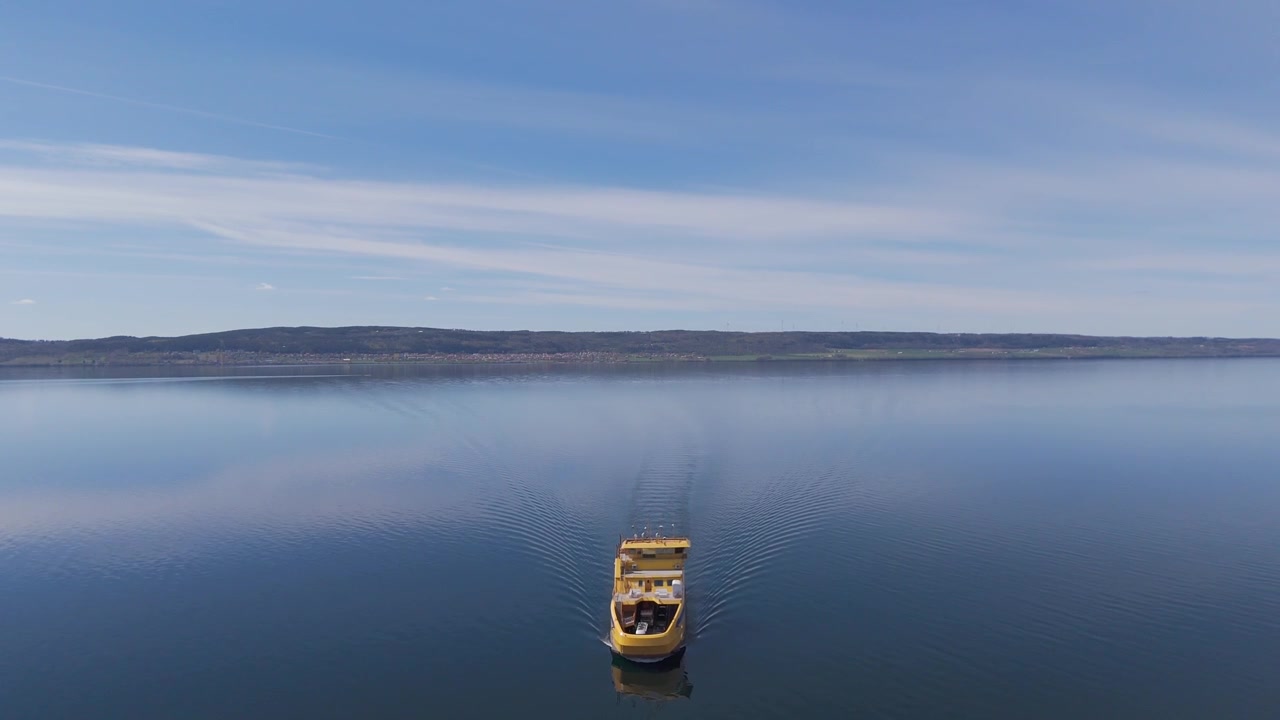 Vista aerea con drone di una barca che naviga su un tranquillo lago scozzese. L'imbarcazione lascia una scia delicata sull'acqua dolce immobile sotto un cielo blu con una costa distante