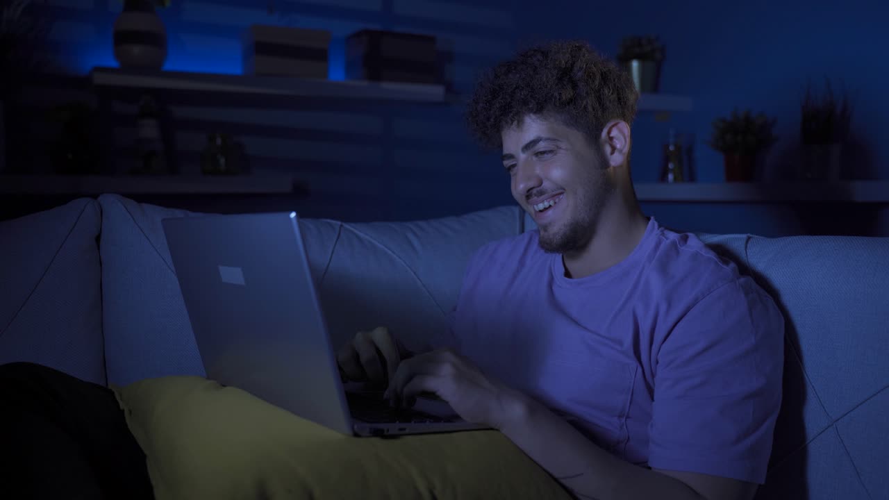 un joven feliz usando una computadora portátil en casa por la noche.
