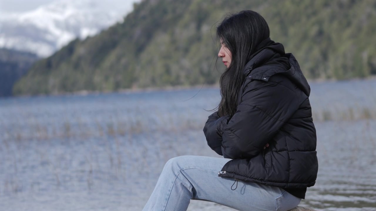 A woman in a black jacket sits pensively by a lakeside, surrounded by snow-dusted mountains. Ideal for themes of solitude or introspection.