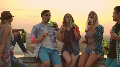 three girls and three boys in plaid shirts drinks beer from green bottels on the party with friends on the roof at the sunset.they are sitting together eat hot pizza after in summer evening.