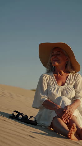 senior woman in white dress and hat relaxing on sand dunes