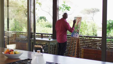 feliz hombre mayor afroamericano de pie en un balcón soleado pintando una imagen de la vista