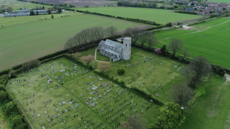 Aerial-Drone-Shot-Flying-Over-Old-Rural-Medieval-11th-Century-Round-Tower-Church-with-Old-Man-Walking-Through-Graveyard-in-North-Norfolk-UK