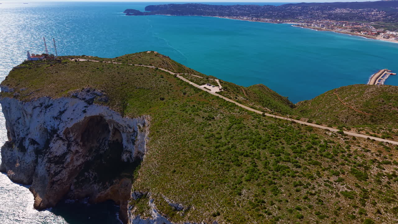 Promontorio costiero roccioso con macchia mediterranea verde, scogliere di calcare bianco, grotta marina, strada tortuosa e faro che si affaccia su una baia mediterranea turchese con una città costiera
