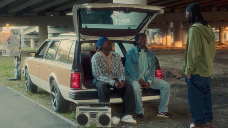 african american friends sitting in open car trunk and talking under overpass