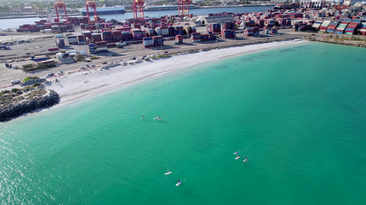 Stand up paddle boarders enjoy the calm waters of Port Beach in Fremantle, Western Australia. The shipping containers of the port are visible in the background, providing a contrast to the beach