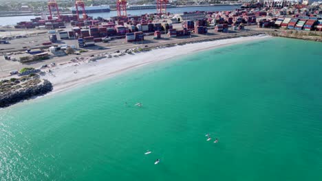 stand up paddle boarders enjoy the calm waters of port beach in fremantle, western australia. the shipping containers of the port are visible in the background, providing a contrast to the beach