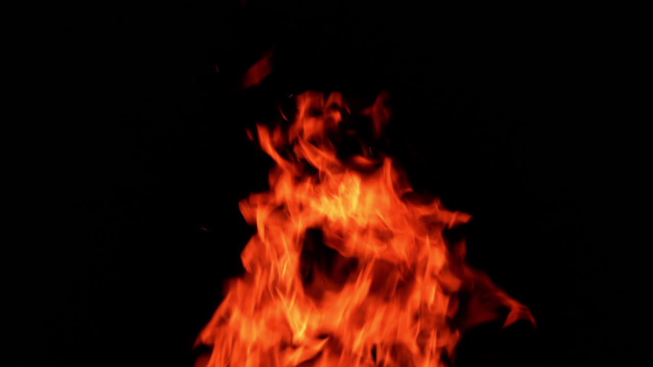 Hot flames of campfire striving up in the air. Multiple ember particles fly in the air at black backdrop.