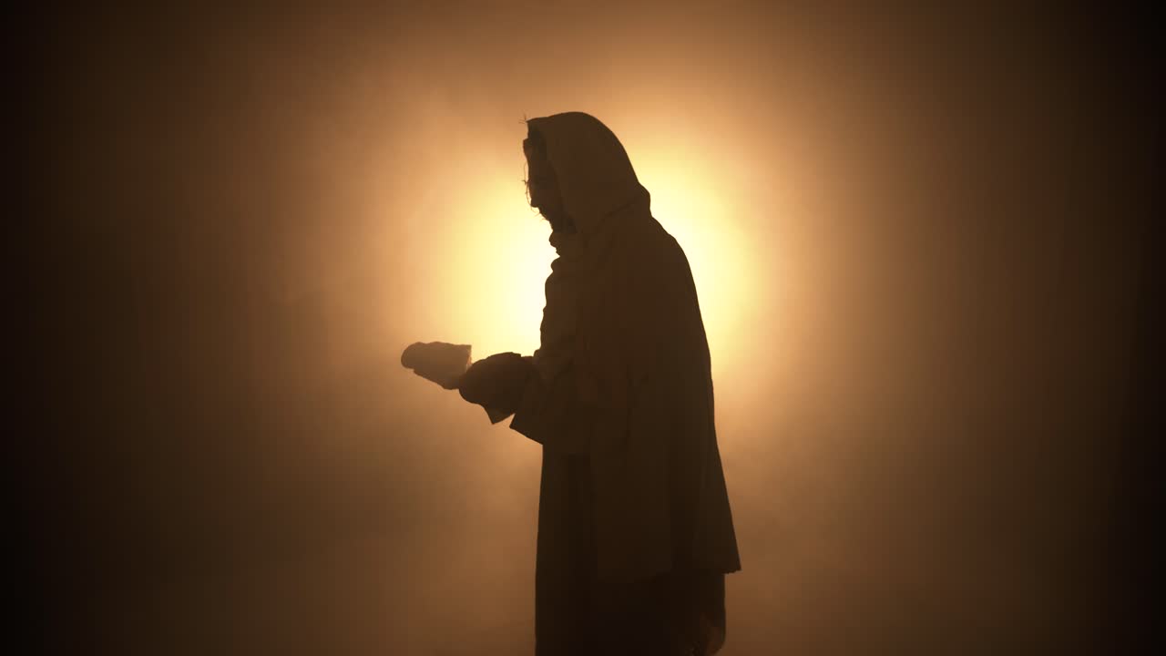Silhouette of Jesus Christ in glowing light, in white hooded robe holding bread as he stands in glowing, bright light praying. Christianity, bible, easter visual.
