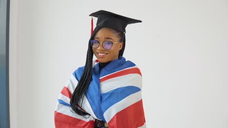 feliz elegante estudiante afroamericana en uniforme de graduado se queda frente a la cámara y sostiene la bandera británica en los hombros
