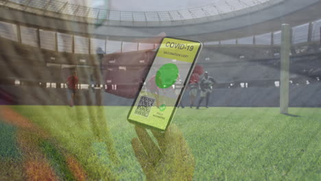 animation of woman holding smartphone with covid 19 vaccination passport over football players