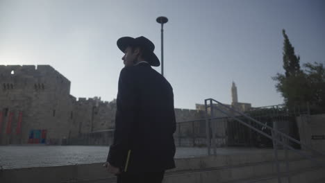 jerusalem, israel, an orthodox jewish man climbs stairs in front of old city and the tower of david walls wearing a black suit and hat and holding a prayer book on a sunny morning