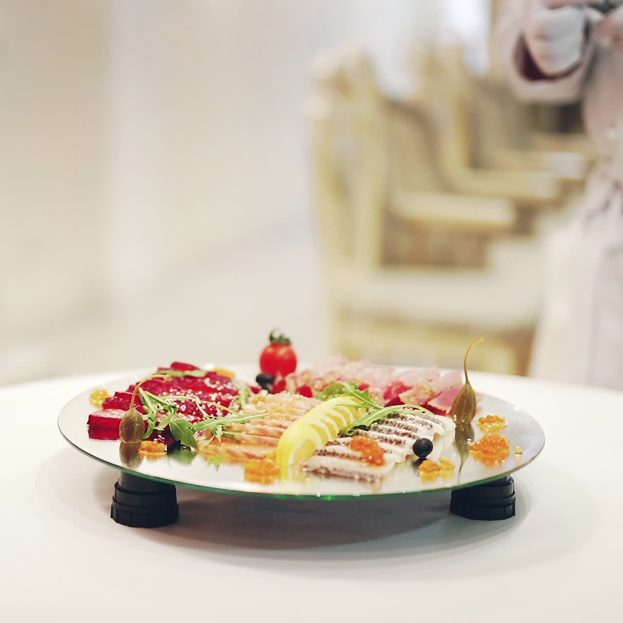 Unrecognizable male cook in white gloves decorates plate with delicious food on a glass turntable. Professional cook puts slicing products prettily for guests.