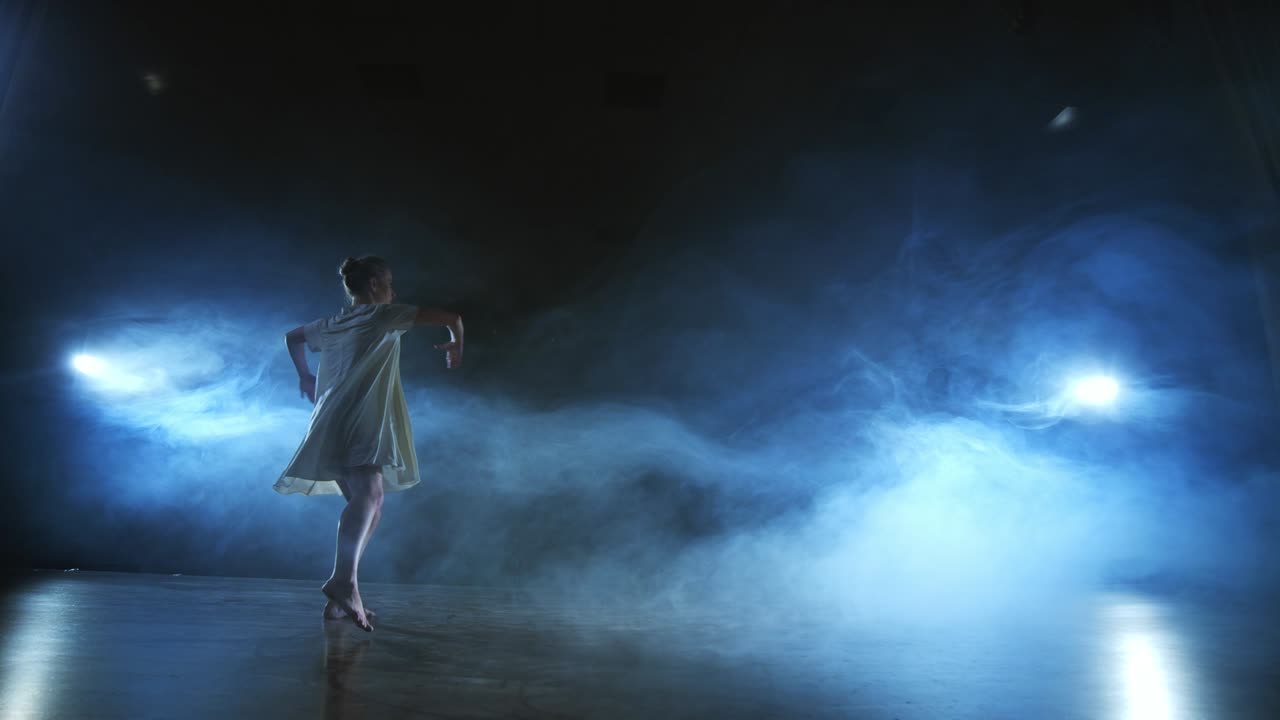Modern ballet dancing woman barefoot doing spins and pirouettes and dance steps standing on stage in smoke in slow motion. Performance on stage