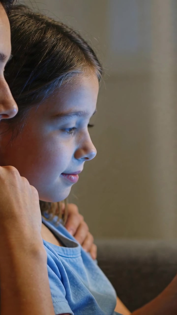 Video verticale: Il bagliore del tablet illumina un bambino con una maglietta azzurra che guarda il tablet sul divano, la mamma gli tiene il braccio.