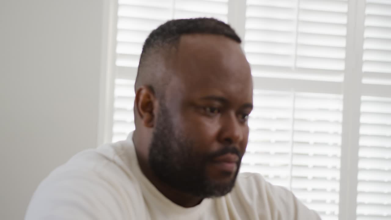 Mid-adult African American man reading screen below while camera focusing at home with blinds